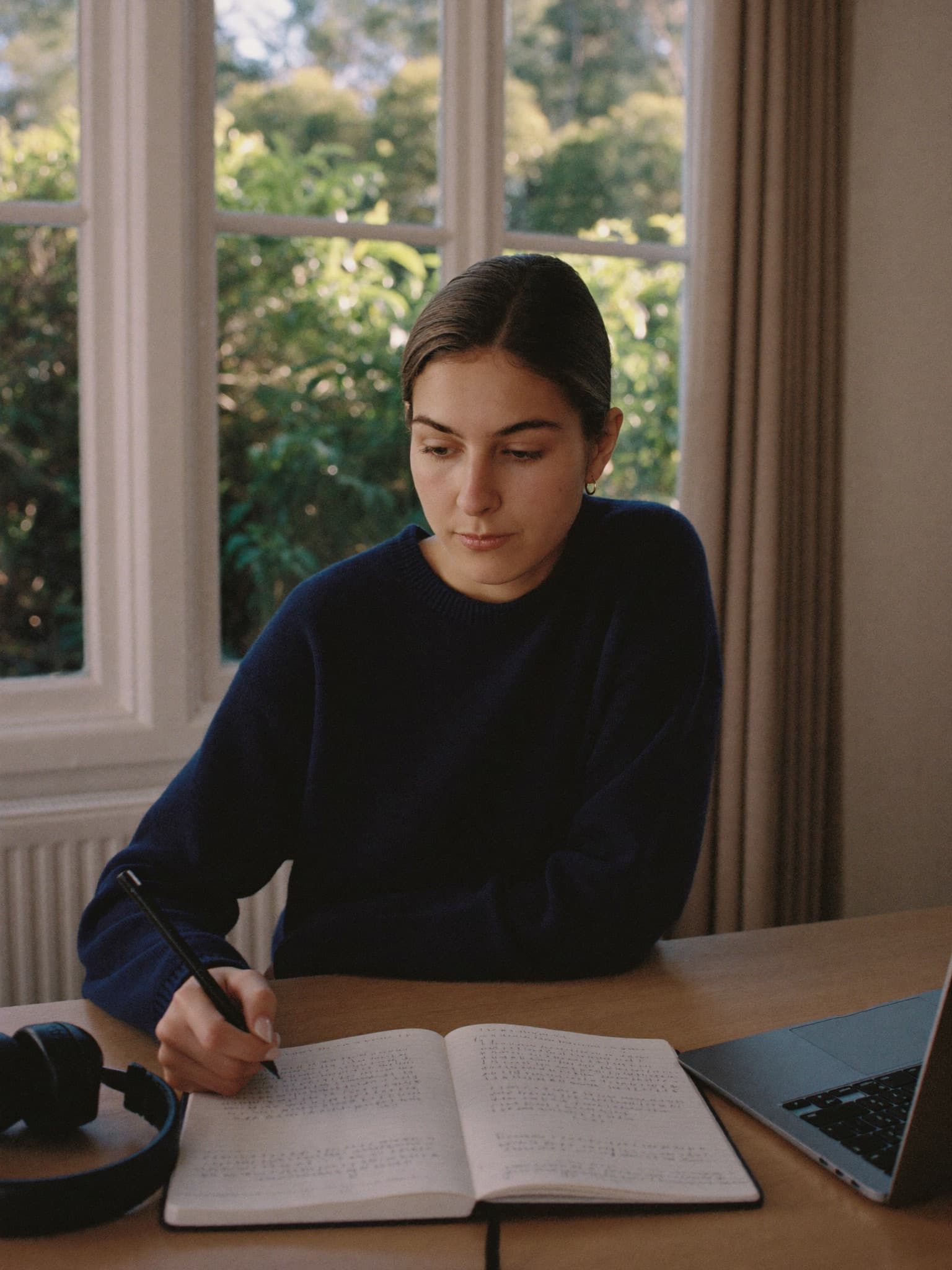 A learner in a navy sweater writing practice notes at a desk by a bright window, laptop and headphones beside her